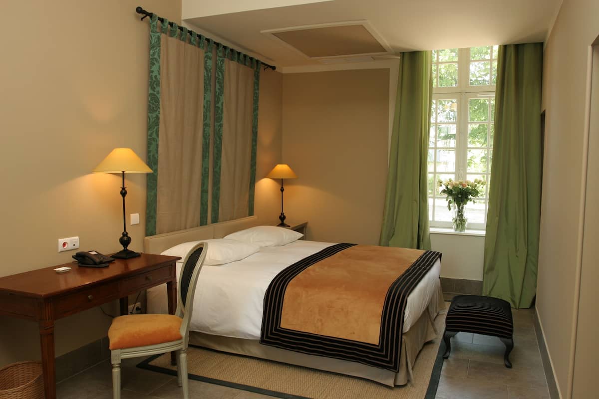 A hotel room with a bed, two bedside lamps, a desk with a chair, and a window with curtains.
