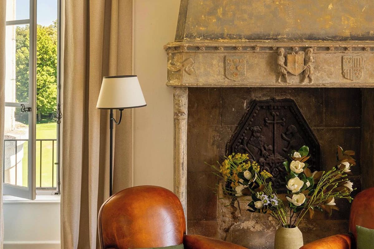 A room with a fireplace, two leather armchairs, a lamp, and a view through a window with curtains.