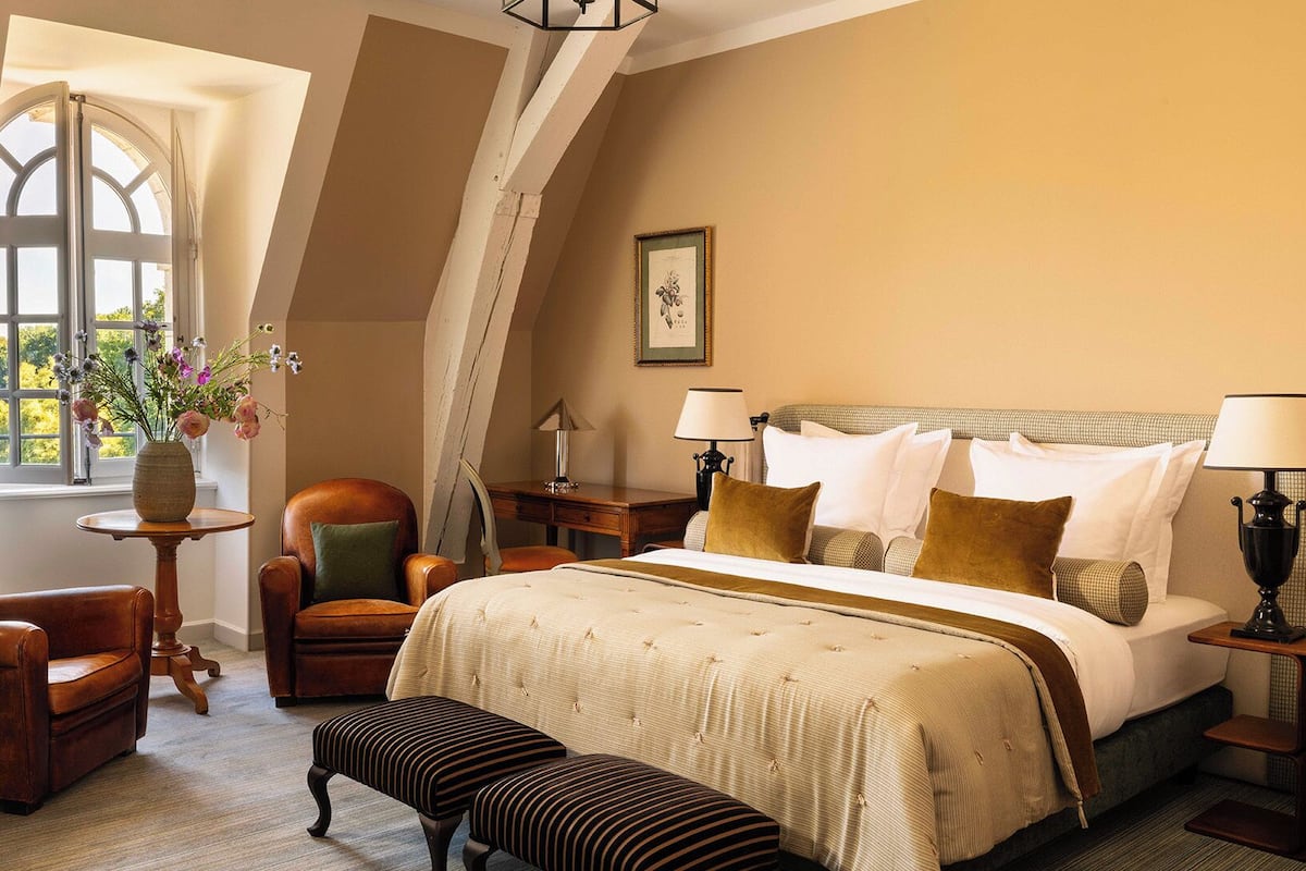 A hotel room with a large bed, two armchairs, a small table with a vase of flowers, a desk, and a window with a view.
