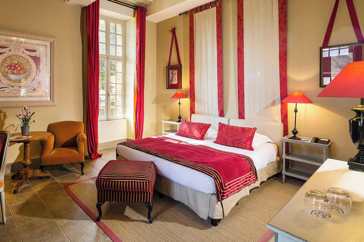 A hotel room with a bed, two bedside tables, a desk, a chair, a window with curtains, and a framed artwork on the wall.
