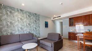 Un moderno salón con un sofá gris, un sillón gris y una pequeña mesa blanca. Se observa una zona de cocina con armarios de madera y una mesa de comedor con sillas.