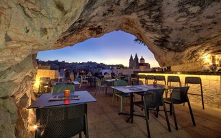 Un espace extérieur aménagé pour les repas, avec des tables et des chaises, une arche en pierre et une vue sur le paysage urbain, avec des bâtiments et une église en arrière-plan.