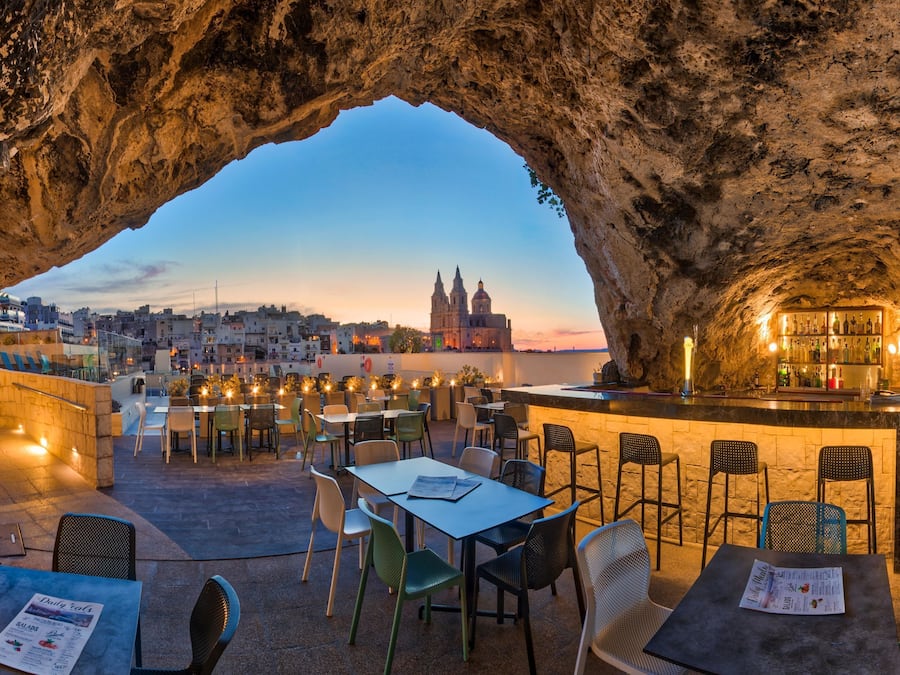 An outdoor dining area with tables and chairs, a bar, and a view of a cityscape through a rock archway.