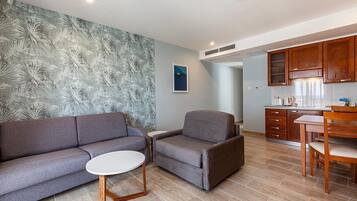 Una sala moderna con un sofá gris, una pequeña mesa redonda y un sillón gris. Se ve, al fondo, una cocina con armarios de madera y una mesa de comedor.