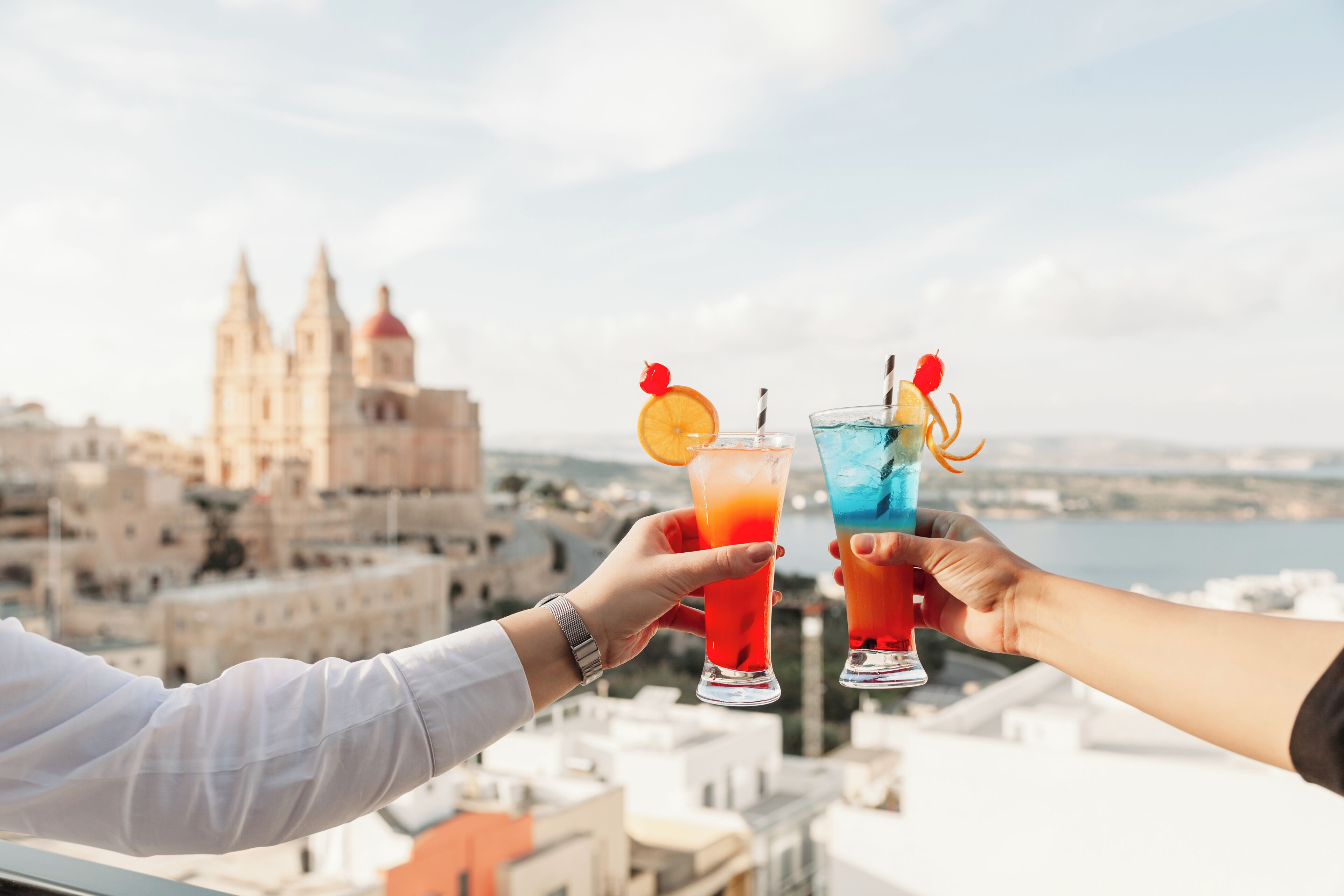 Two people toasting with drinks, a cityscape with a prominent cathedral in the background, and a body of water.