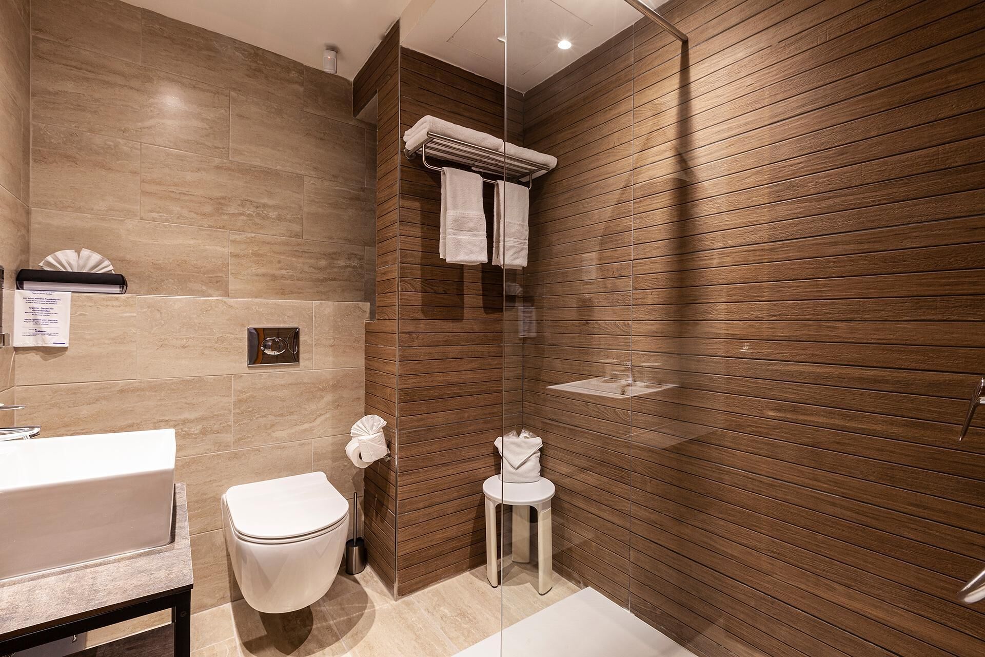 A modern bathroom with a wooden panel wall, a white toilet, a glass shower enclosure, and a white sink.