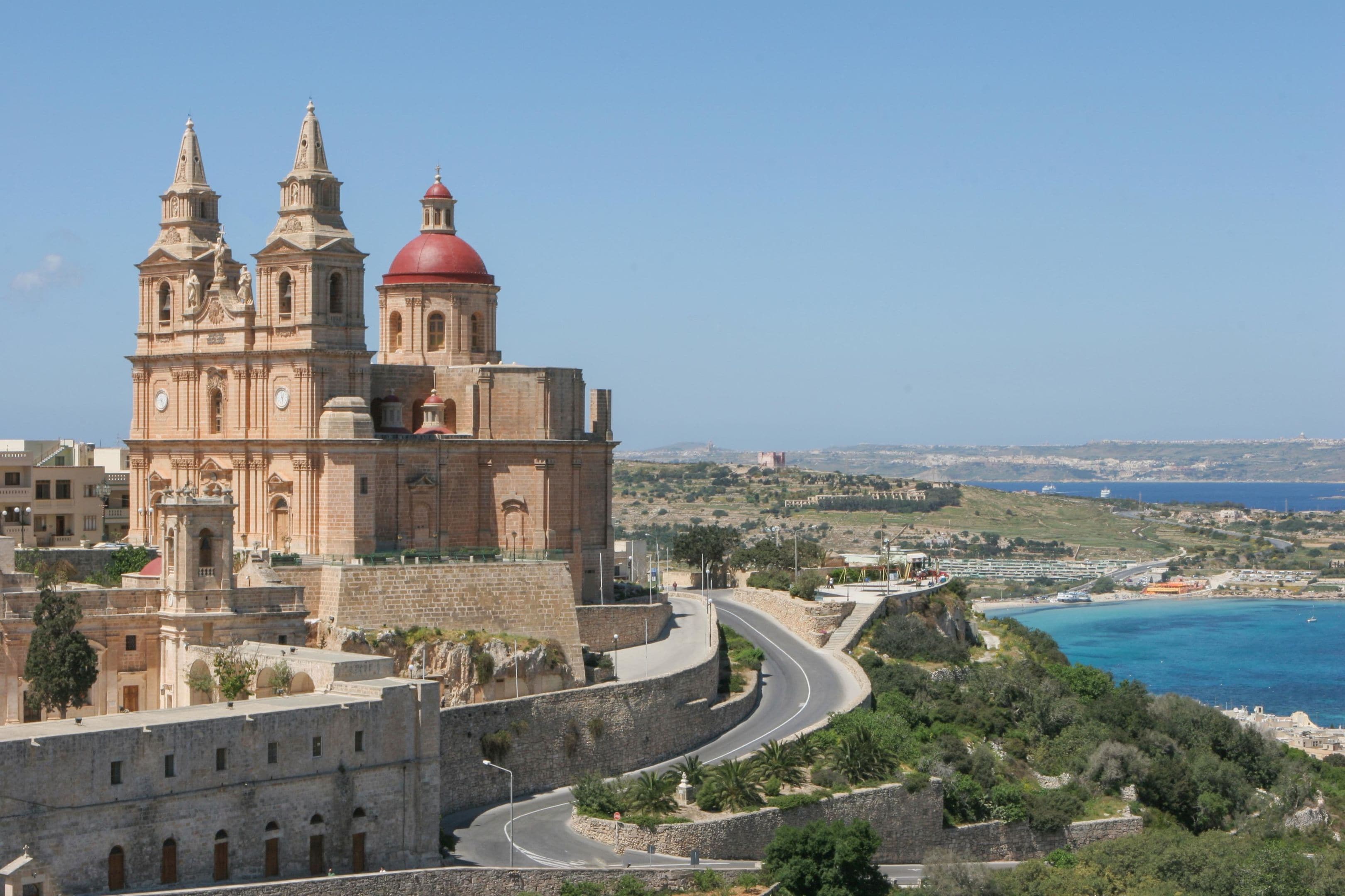 A historic church with two towers and a red dome, surrounded by a walled city with a curved road and coastal views.