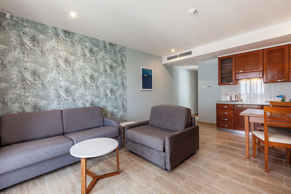 A modern living room with a gray sofa, a gray armchair, and a small white table. There is a kitchen area with wooden cabinets and a dining table with chairs.