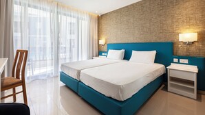 Habitación de hotel con dos camas, una silla de madera, una mesita pequeña y un ventanal con cortinas traslúcidas.