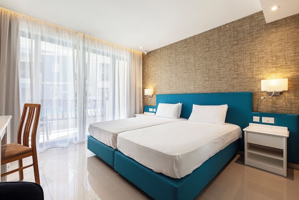 A hotel room with two beds, a wooden chair, a small table, and a large window with sheer curtains. - Pergola Hotel & Spa (Mellieha)