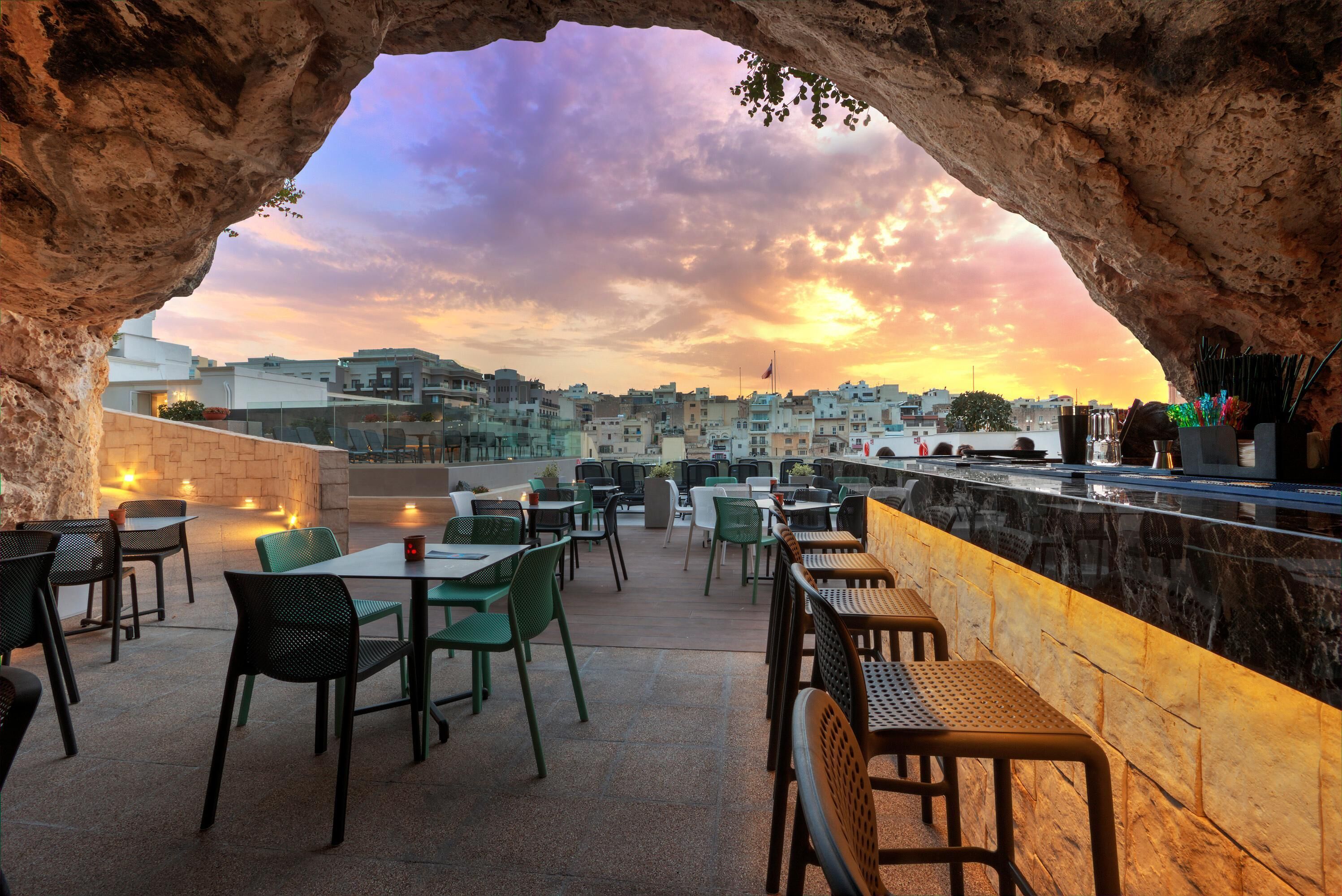 An outdoor dining area with tables and chairs, a stone wall, and a view of buildings under a sunset sky.