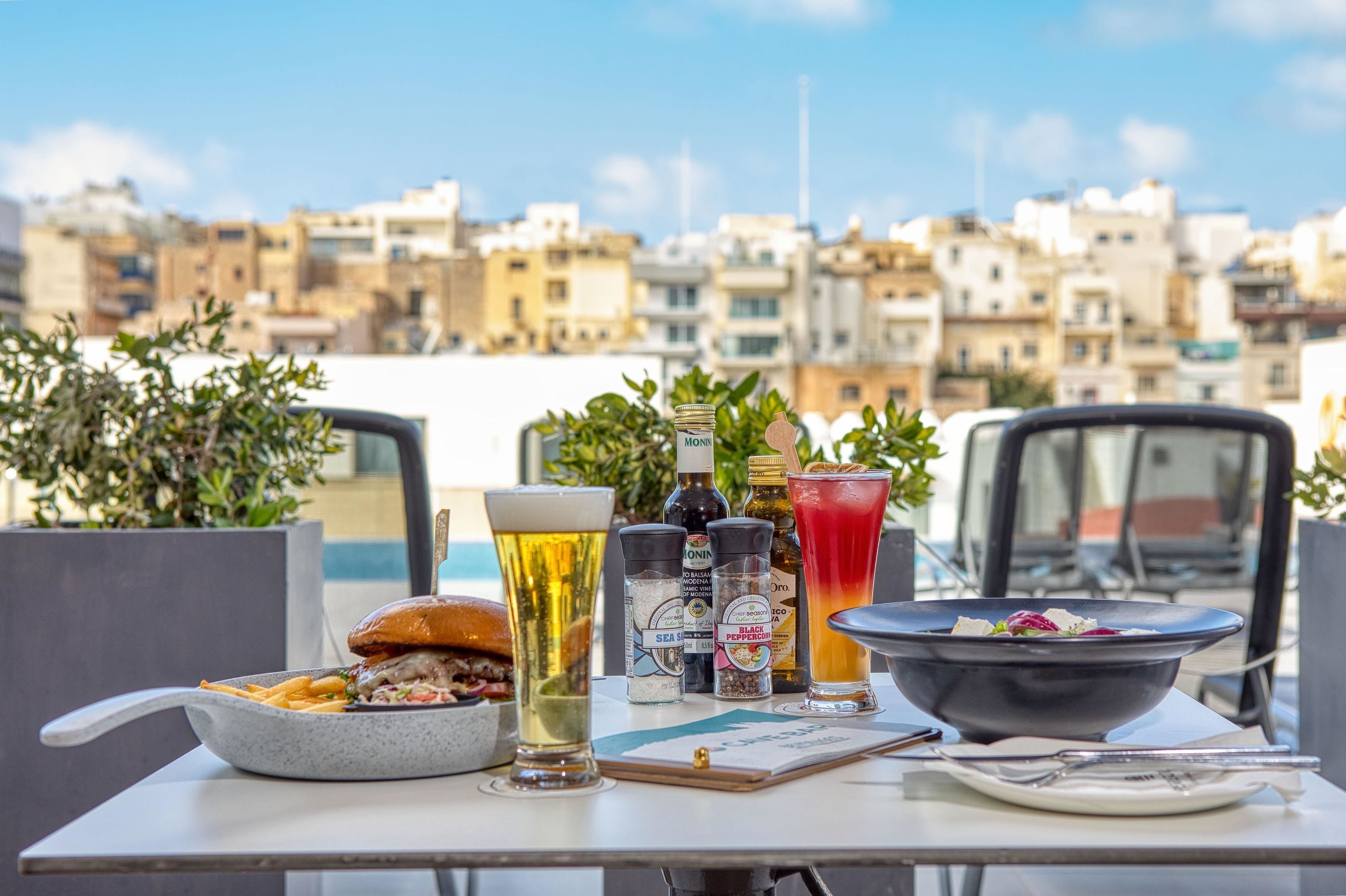 Une table avec un hamburger, une bière, une salade, des condiments et un menu, le tout installé sur un toit offrant une vue sur la ville.