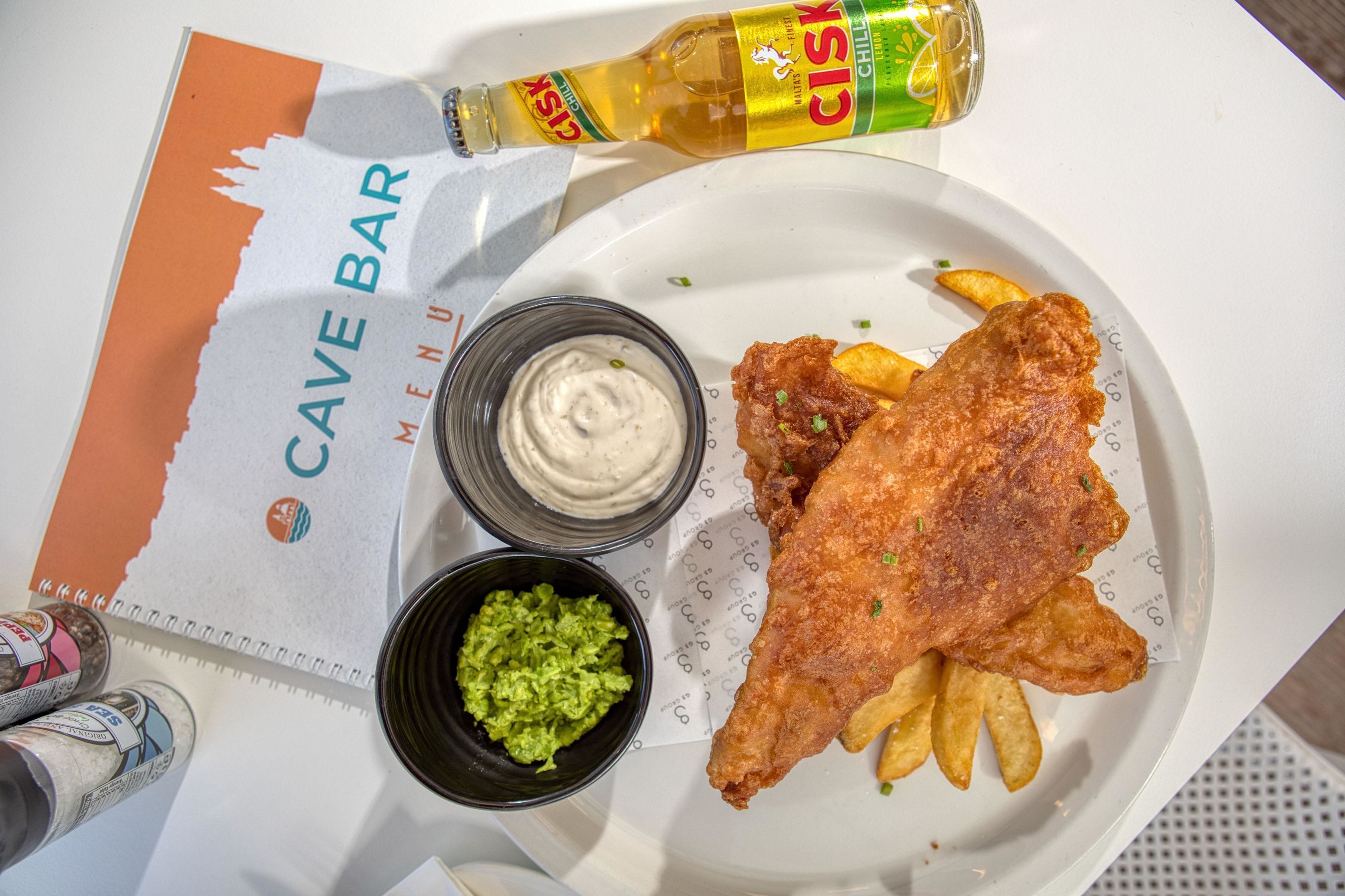 Een maaltijd met vis en friet, een biertje en sauzen op een tafel.