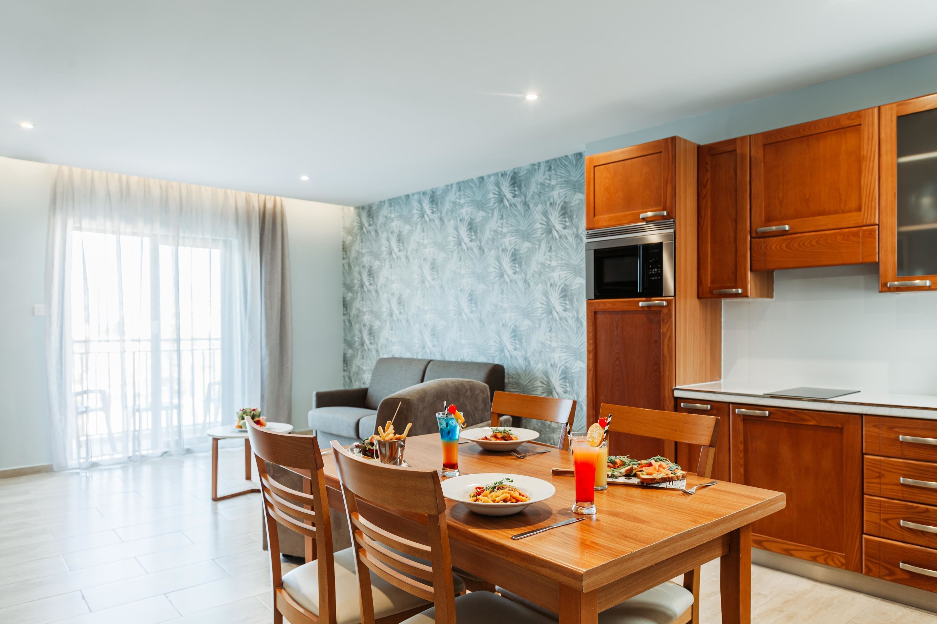 Una cocina moderna con armarios de madera, una mesa de comedor para dos personas y un ventanal con cortinas traslúcidas.