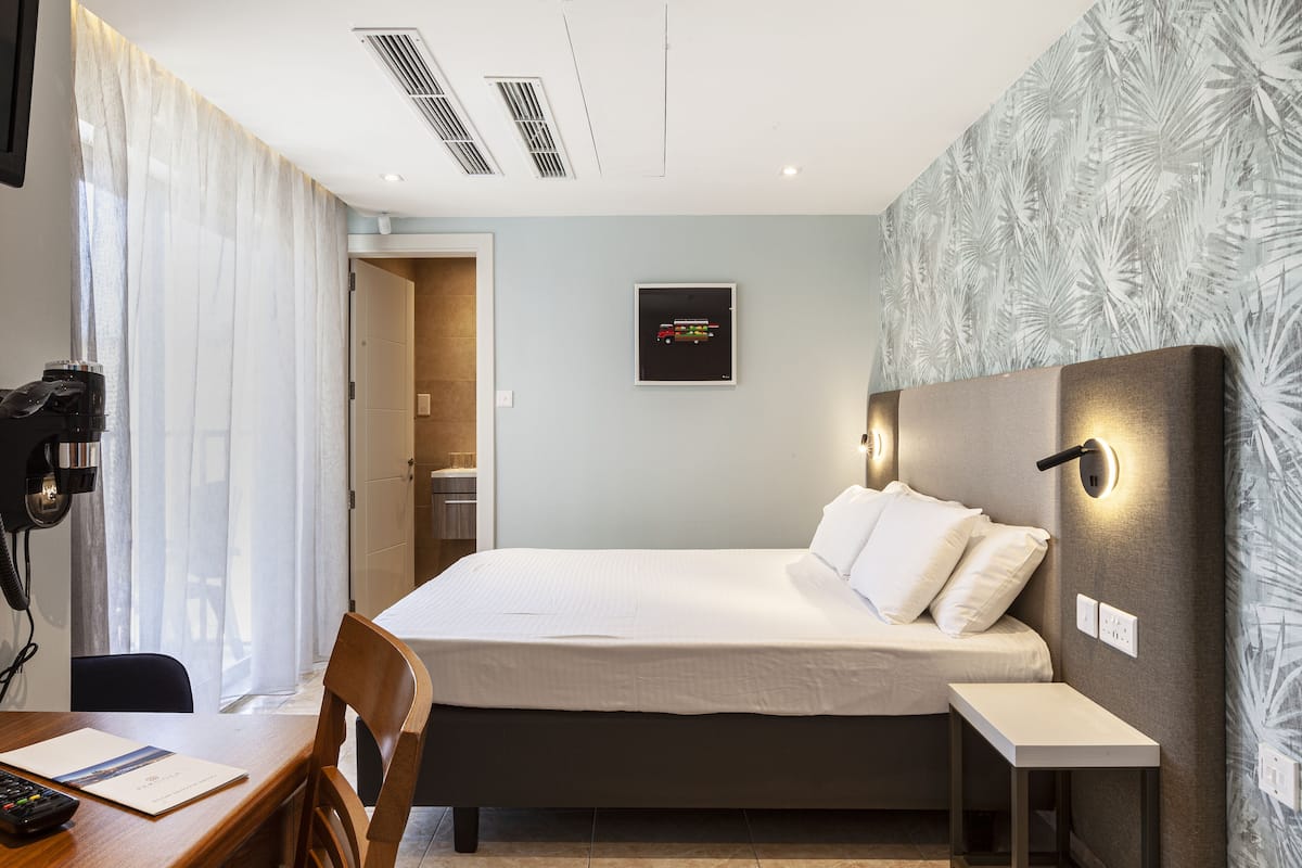 A hotel room with a bed, bedside table, chair, desk with a phone, and a wall-mounted TV.