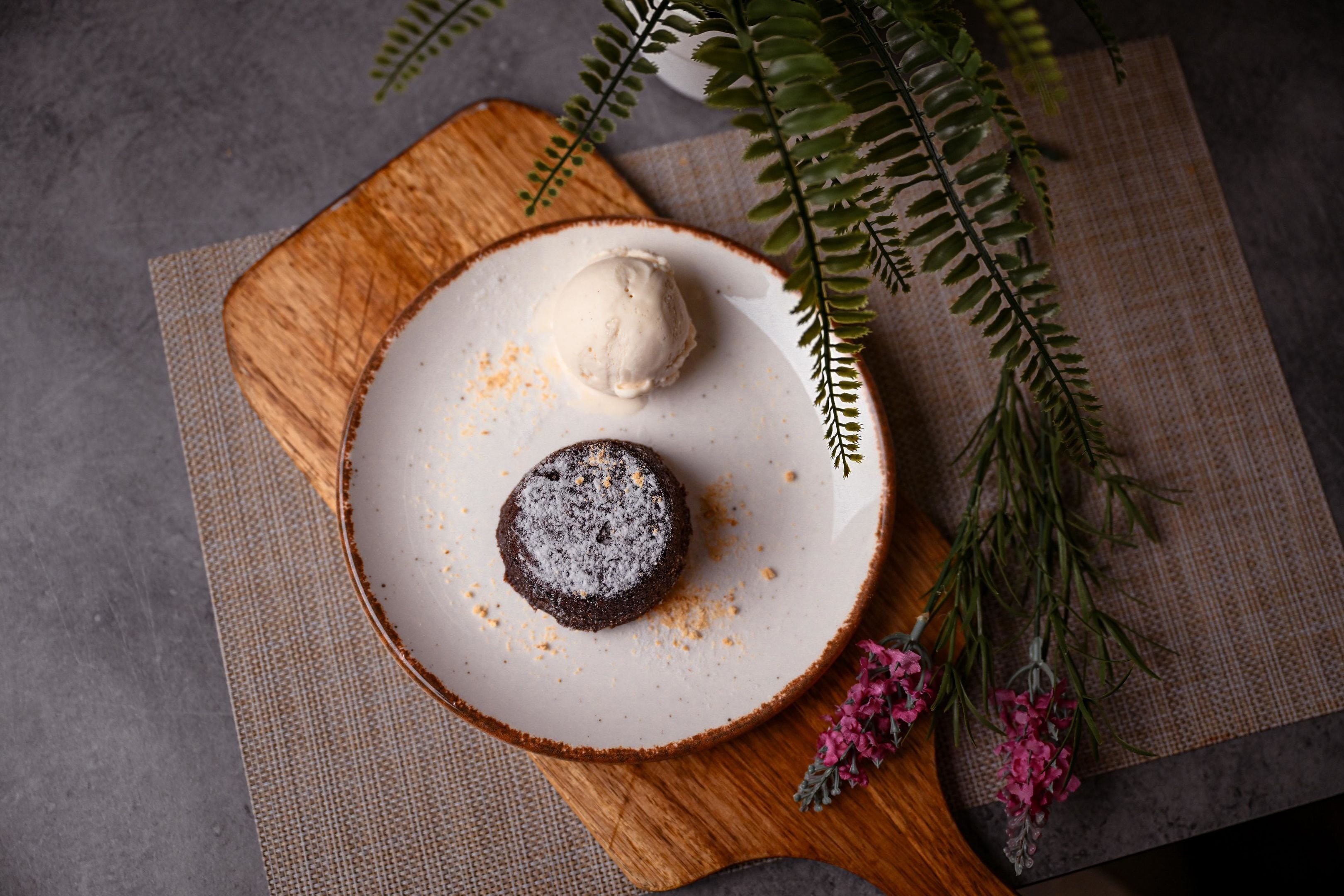 A dessert plate with a chocolate cake and ice cream on a wooden board, accompanied by a fern and pink flowers.