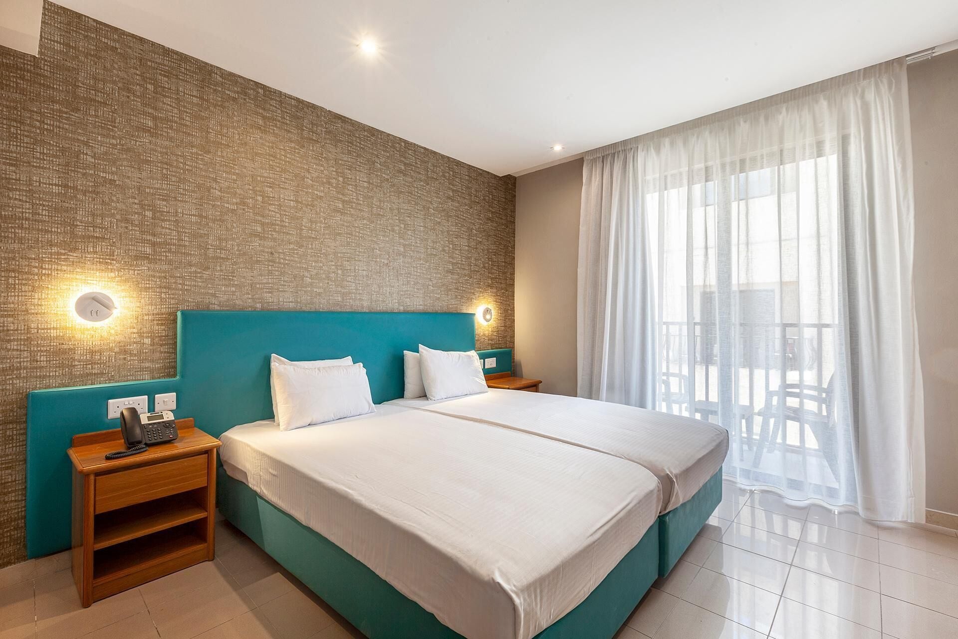 A hotel room with a large bed, two bedside tables, a wall-mounted phone, and a view of the outdoors through sheer curtains.