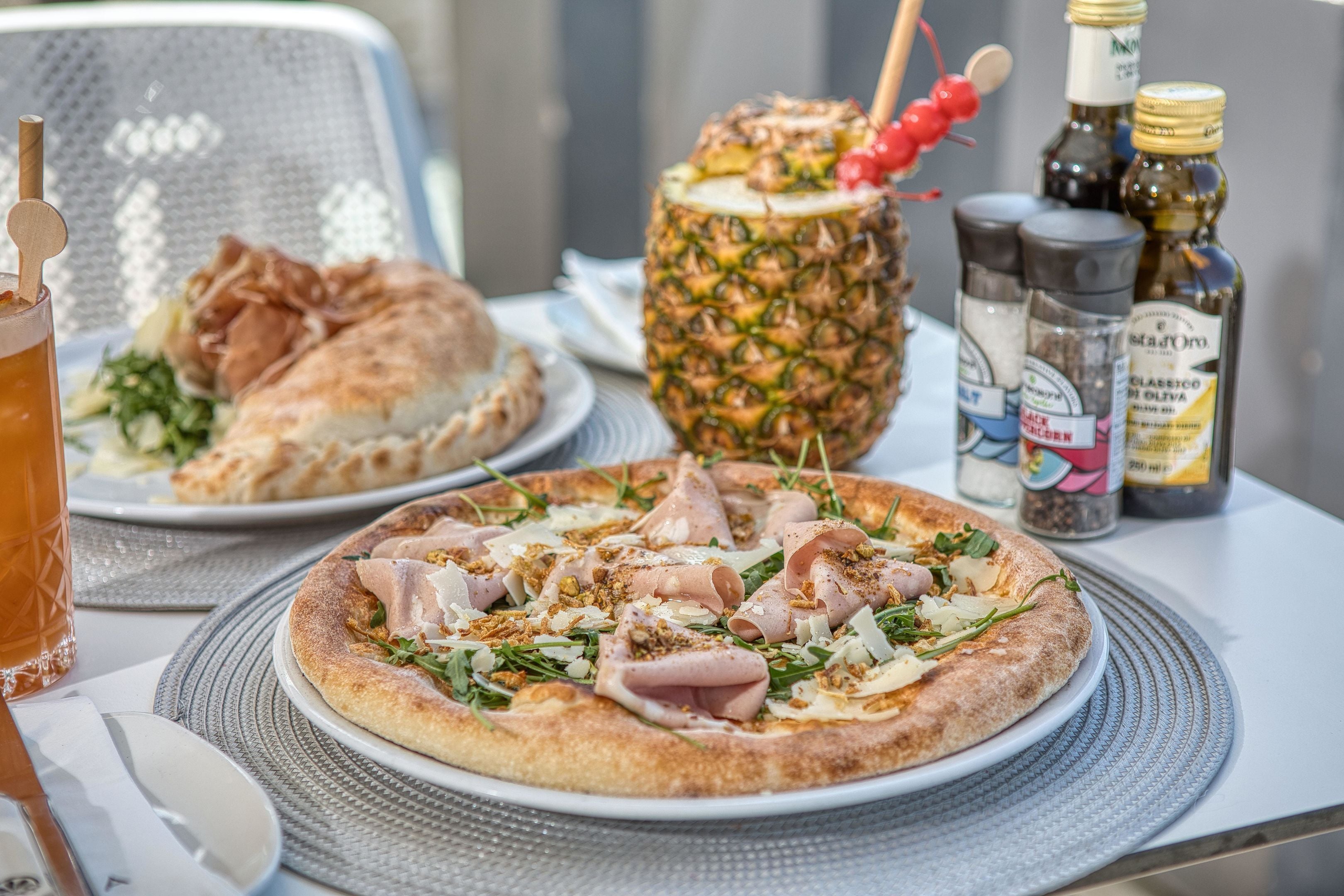 Pizza met diverse toppings op een bord, een ananas aan een spiesje, en sauzen op een tafel.