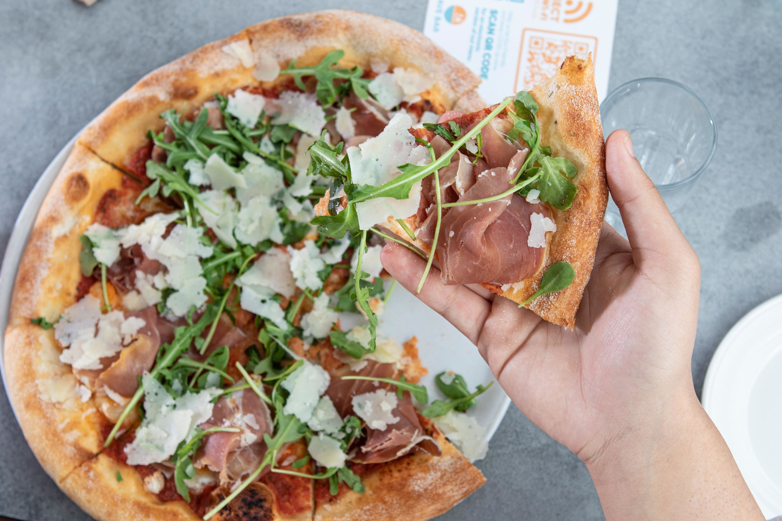 Eine Hand hält ein Stück Pizza mit Rucola und Prosciutto.