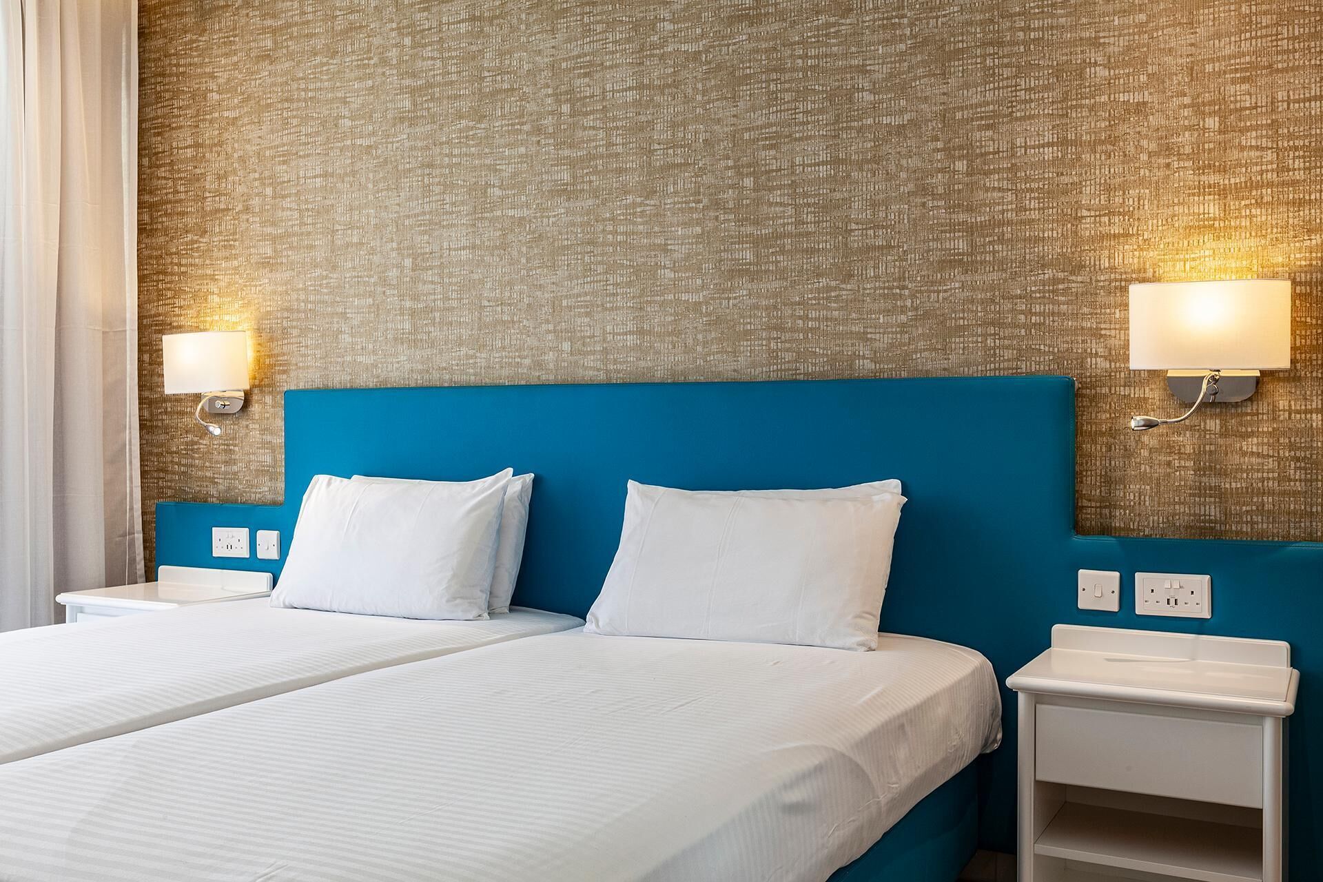 A hotel room with two beds, a blue headboard, and wall-mounted lights.