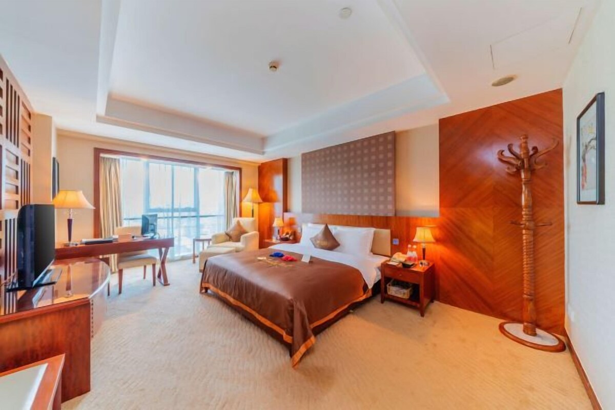 A hotel room with a bed, desk, chair, lamp, and a large window.