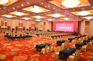 Un amplio salón de conferencias con filas de mesas y sillas, un escenario con una pantalla y una alfombra roja.