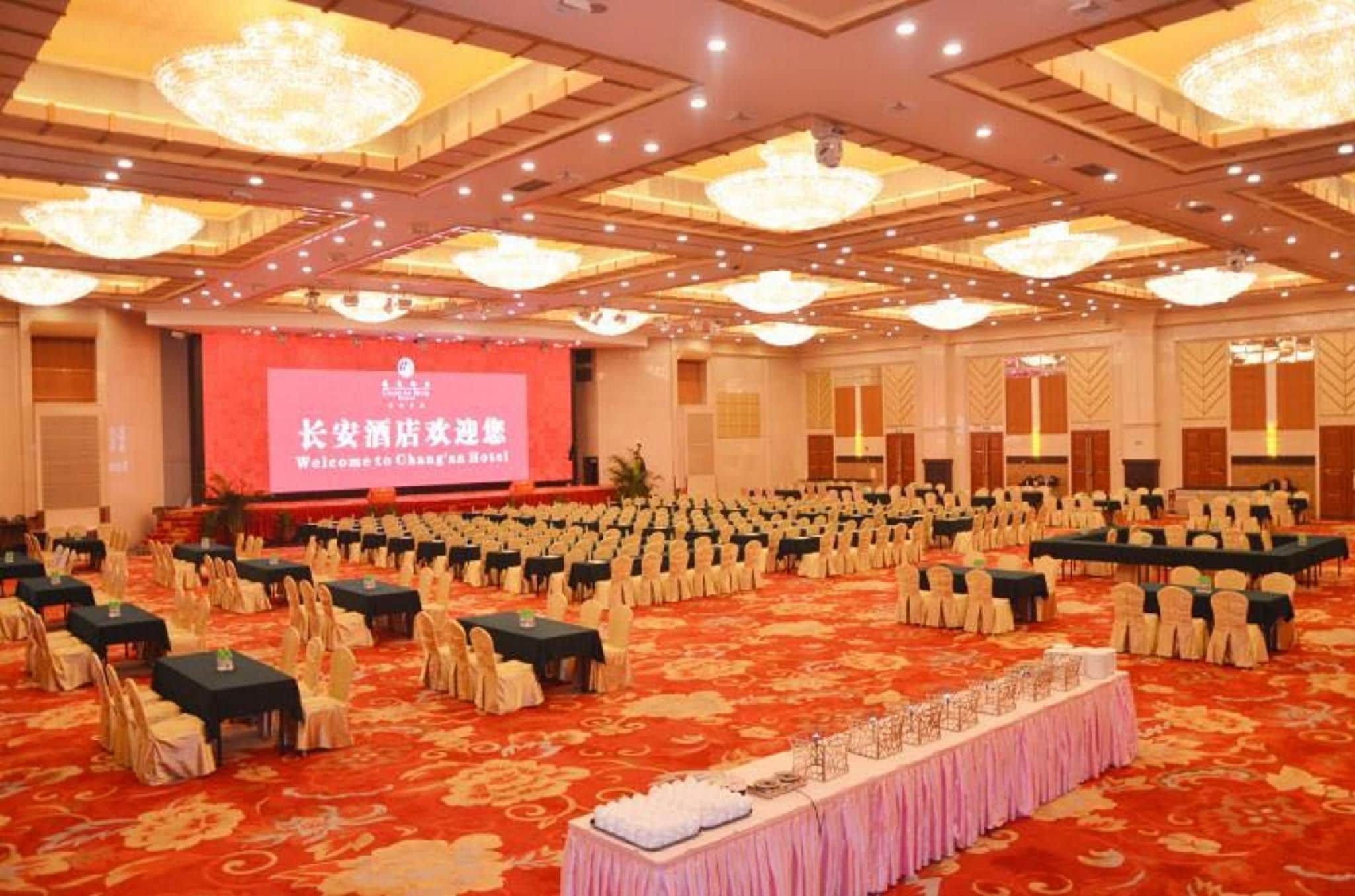 A large conference room with rows of tables and chairs, a stage with a screen, and chandeliers.