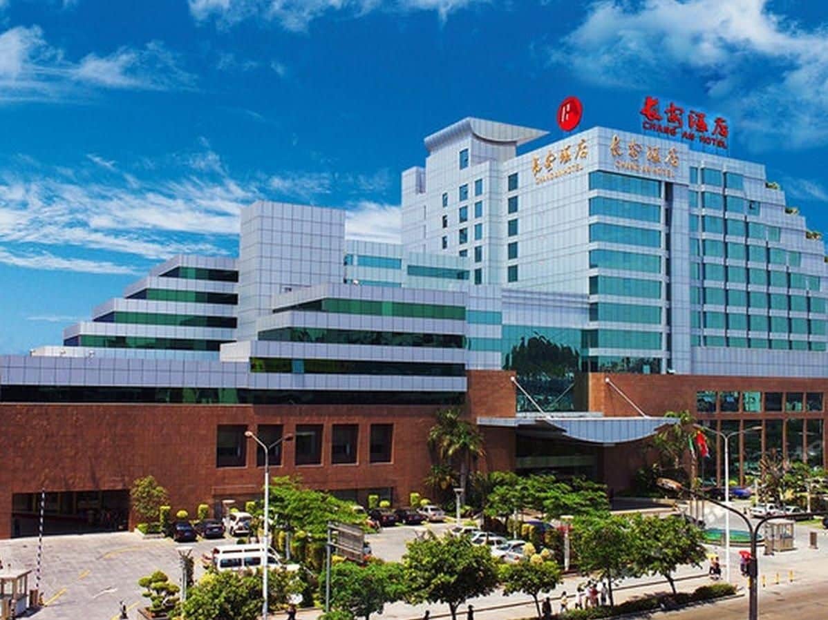 A modern hotel with a prominent red logo, multiple floors, and a parking area with trees.