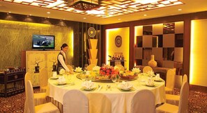 A hotel dining room with a round table set for a meal, a television on the wall, and a service staff member. - Chang an Hotel (Dongguan)