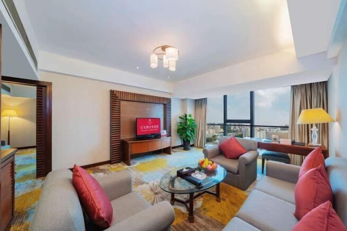 A modern hotel room with a sofa set, a coffee table, a TV, and a large window offering a city view.