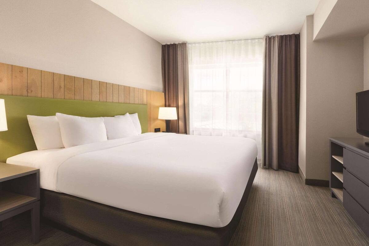 A hotel room with a large bed, a wooden headboard, a nightstand with a lamp, a dresser, and a television.