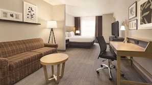 A hotel room with a plaid sofa, a round table, a bed, a desk with a lamp, and framed artwork on the walls. - Country Inn & Suites by Radisson, Conyers, GA (Conyers)