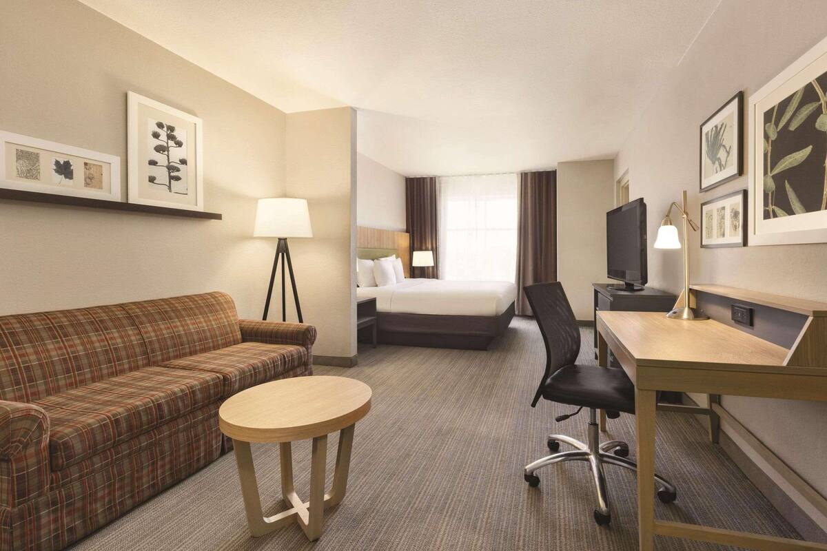 A hotel room with a plaid sofa, a round table, a bed, a desk with a lamp, and framed artwork on the walls.