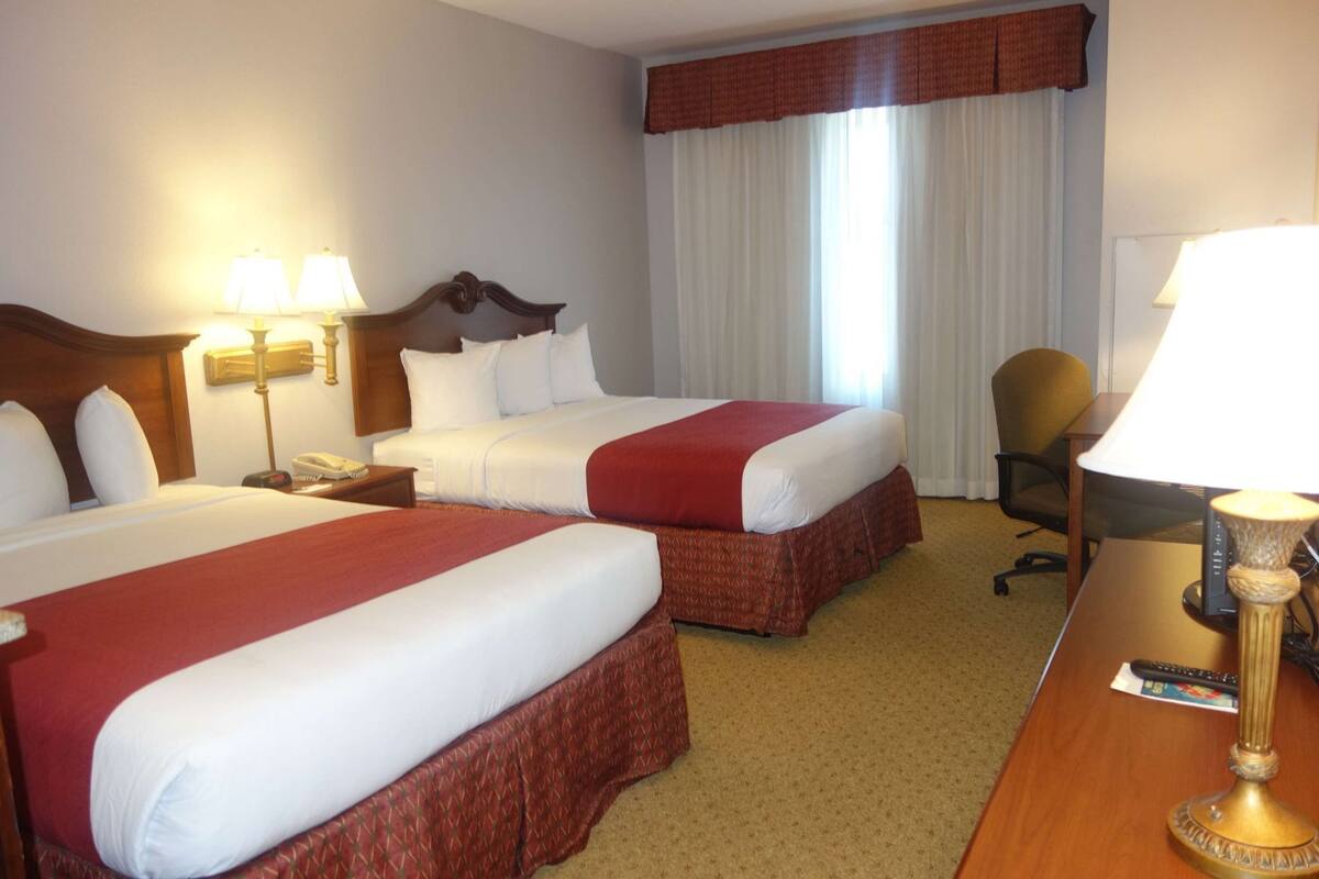 A hotel room with two beds, a desk, a chair, lamps, and a window with curtains.