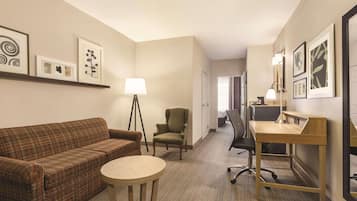 A hotel room with a plaid sofa, a small round table, a desk with a lamp, a chair, and framed artwork on the walls.