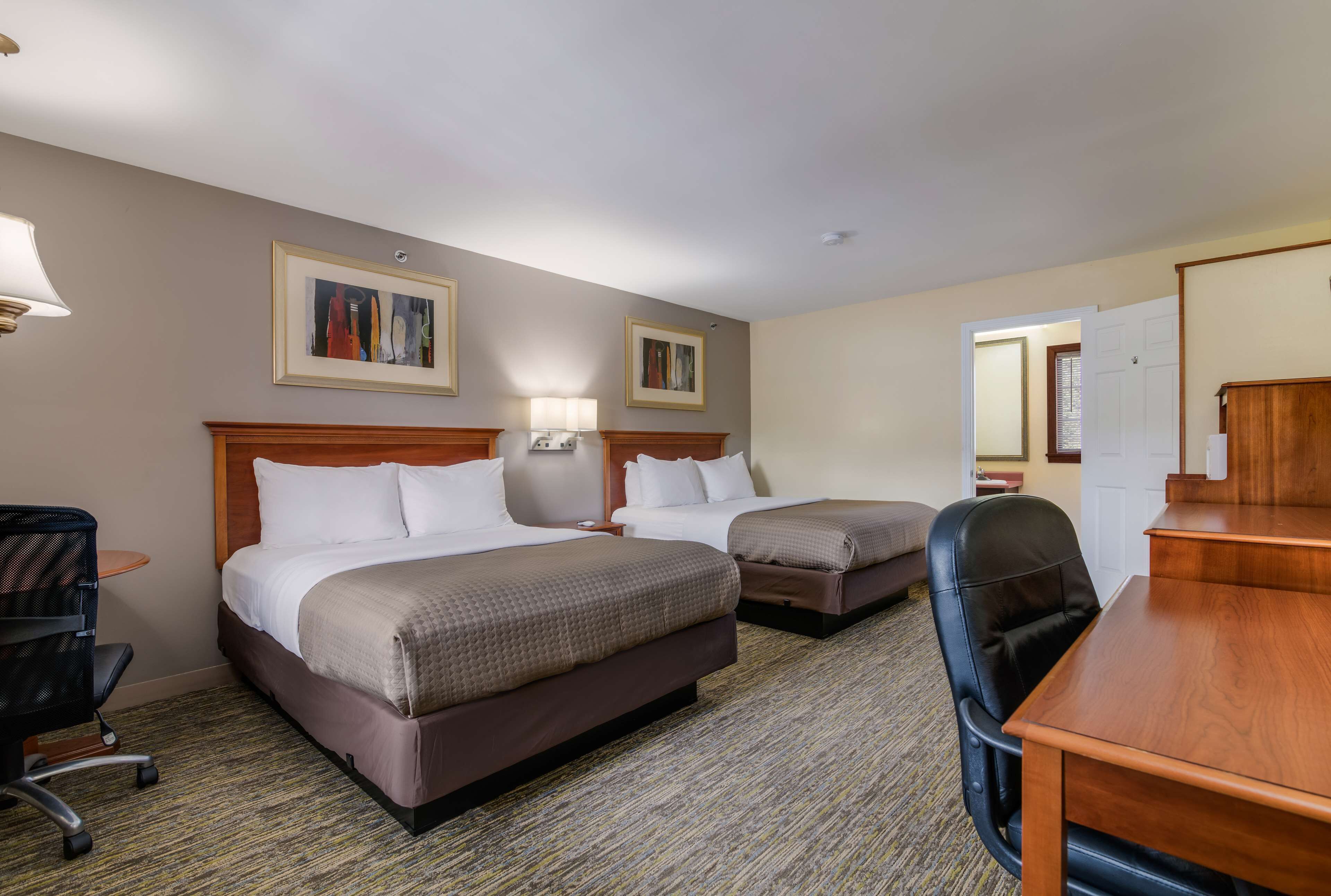 Quarto de hotel com duas camas, uma escrivaninha, uma cadeira, uma luminária e duas fotografias emolduradas na parede.