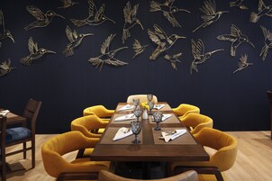 Un comedor con una pared oscura que presenta dibujos lineales dorados de aves volando, una mesa de madera dispuesta con cristalería y sillas tapizadas en amarillo.