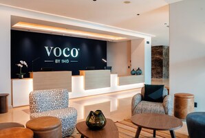 Zona de recepción con fondo negro que muestra la marca 'VOCO by IHG', un mostrador de recepción, asientos con diseño y un jarrón decorativo.
