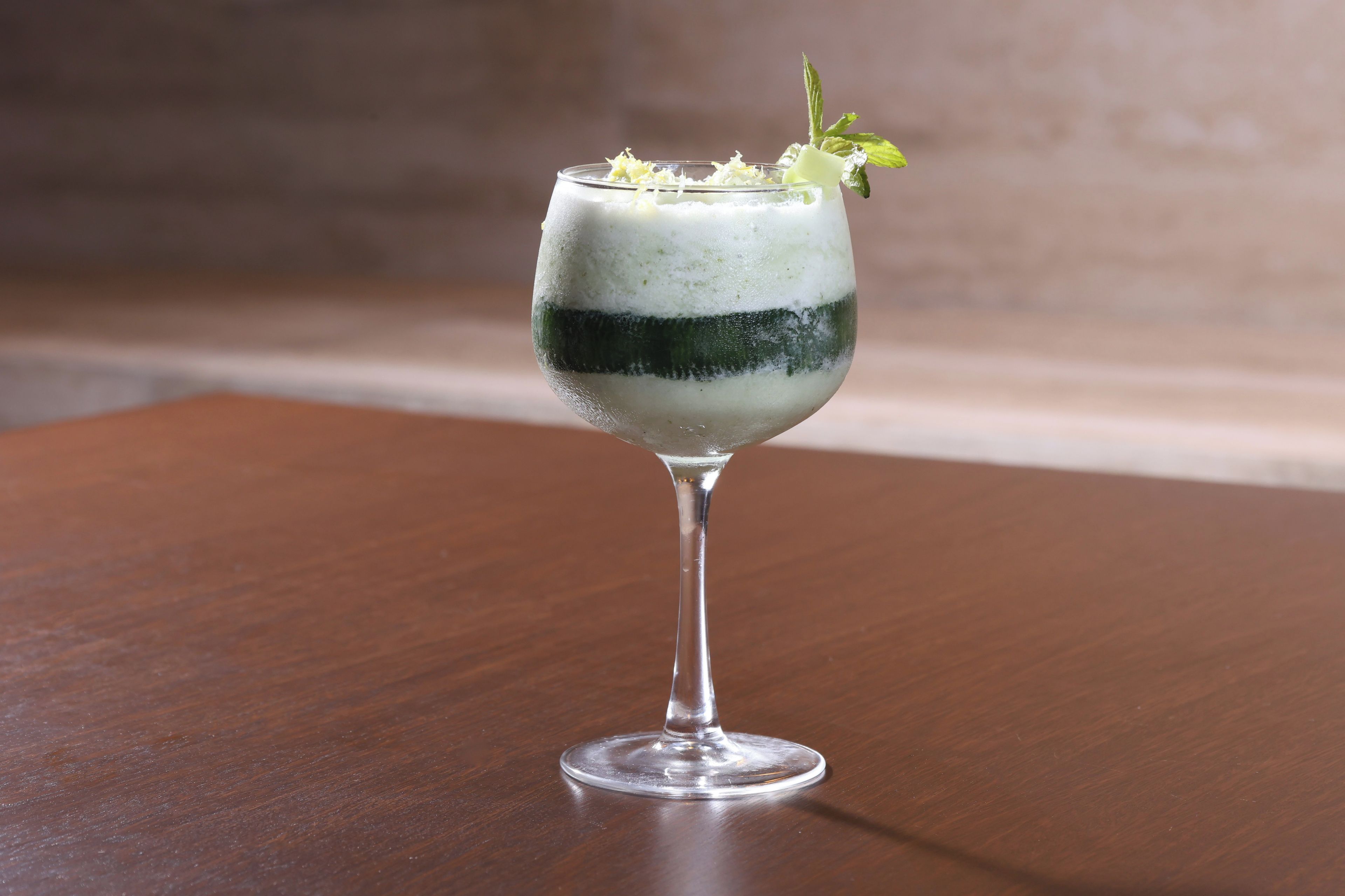 Ein Cocktail im Stielglas mit schichtweise angeordnetem Getränk, garniert mit Minzblättern und Zitronenzesten, auf einem Holztisch.