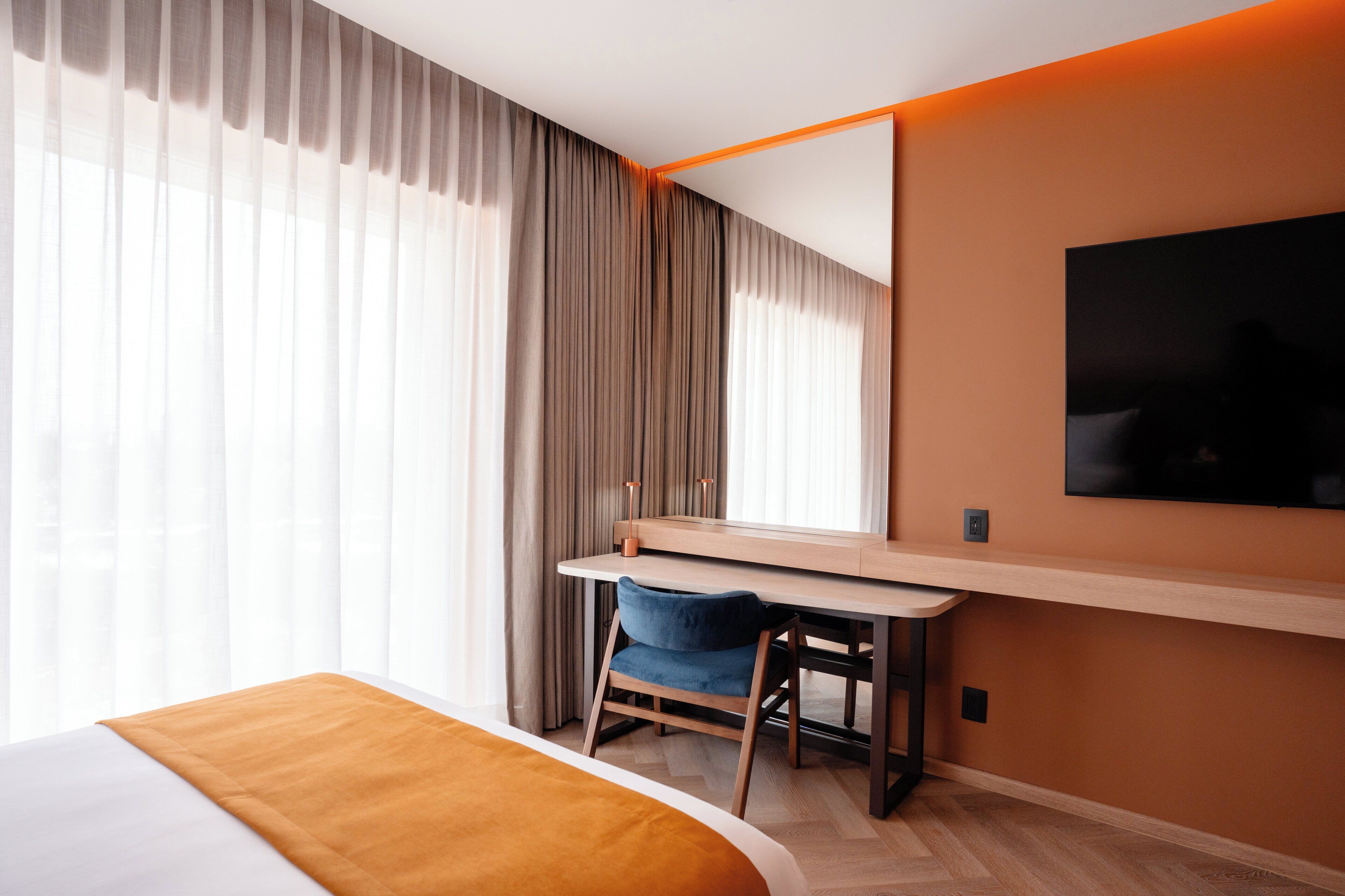 Habitación de hotel con cama, escritorio, silla, televisor y una ventana grande con cortinas.