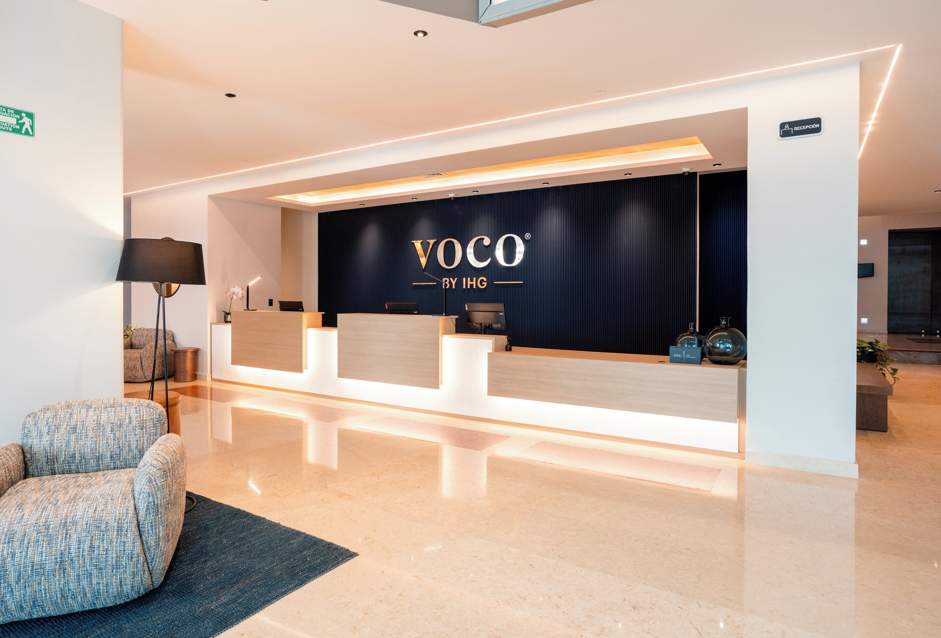 Un espace d’accueil doté d’un fond noir où figure le logo « VOCO BY IHG », un comptoir d’accueil et un coin salon avec une lampe et une plante.