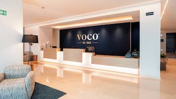 A reception area with a black backdrop displaying 'VOCO BY IHG' branding, a reception desk, and a seating area with a lamp and a plant.