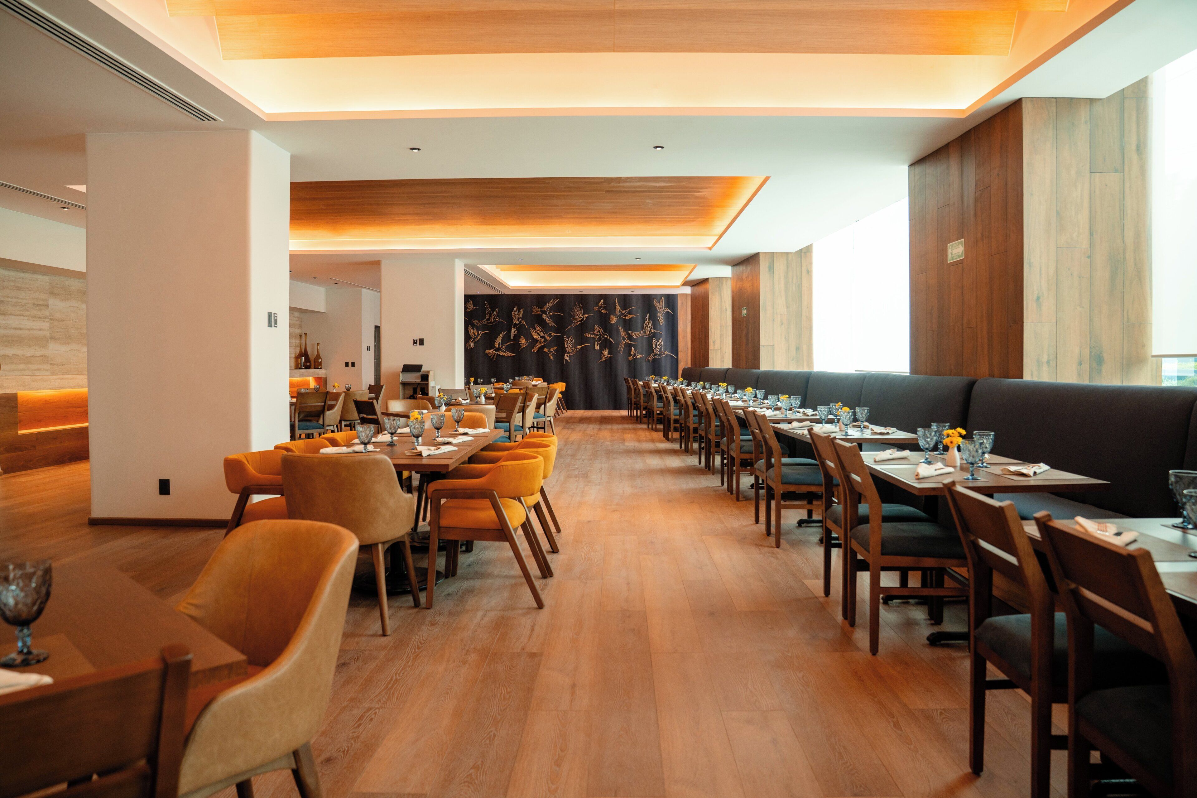A modern dining area with long tables, wooden chairs, and a central wall with a bird mural.