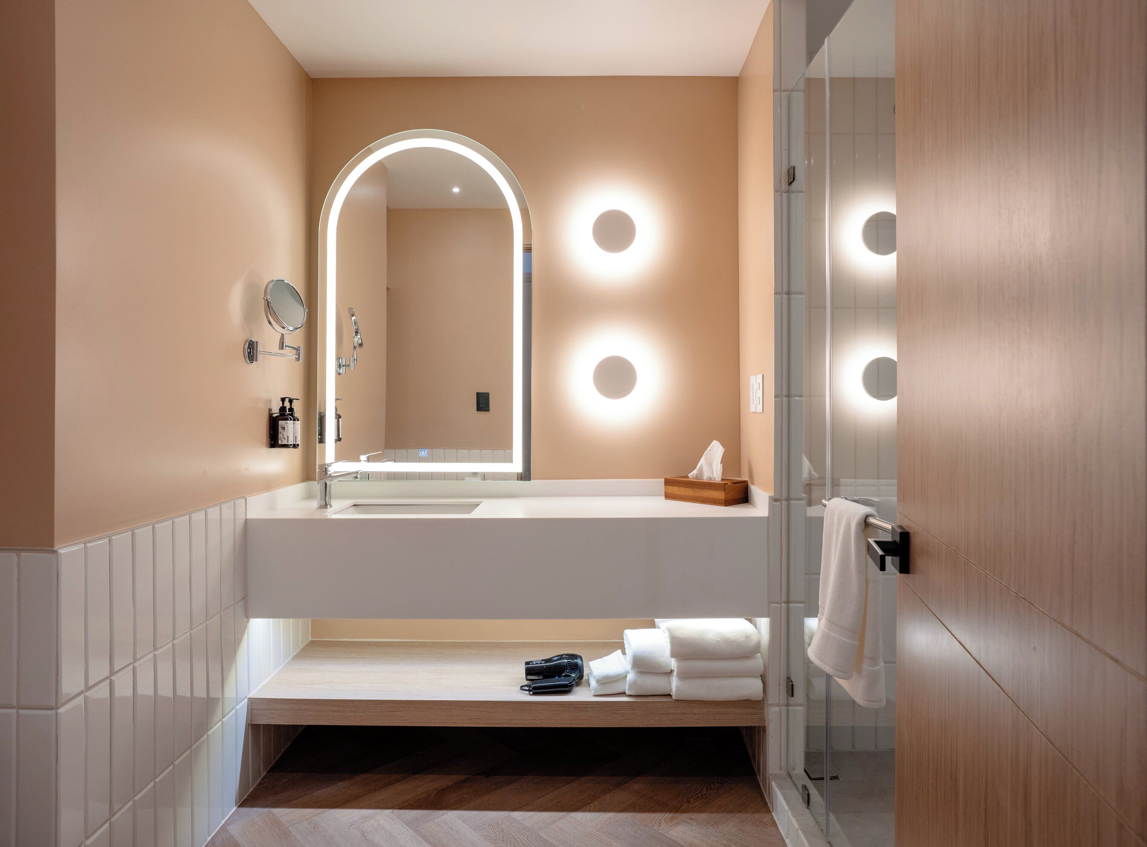 Un baño moderno con un espejo grande, luces redondas en la pared, un tocador de madera y un box de ducha de cristal.