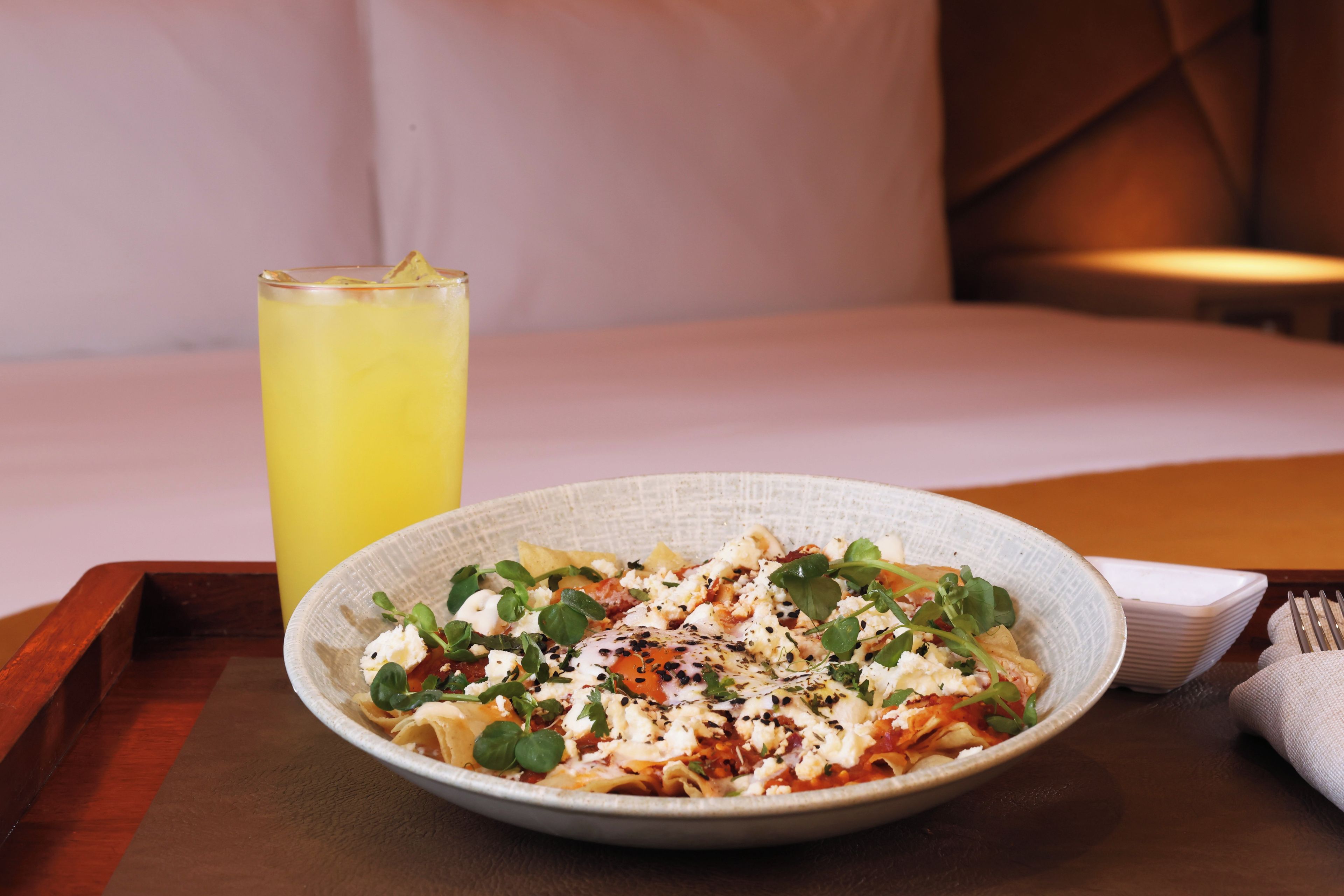 A plate of pasta with greens and cheese, a glass of yellow drink, and a fork on a wooden tray.