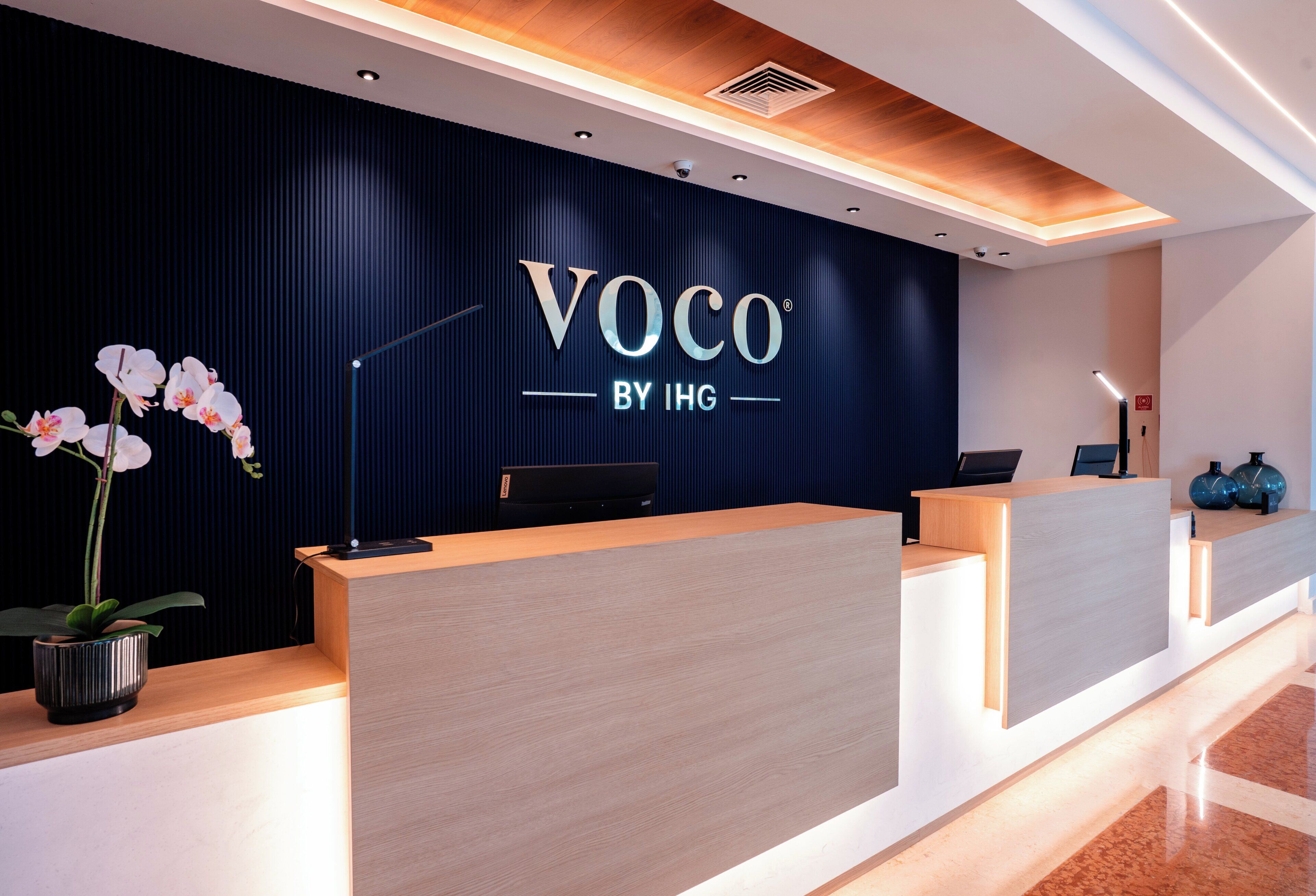 Una recepción con una pared oscura y el texto 'VOCO BY IHG' de fondo.