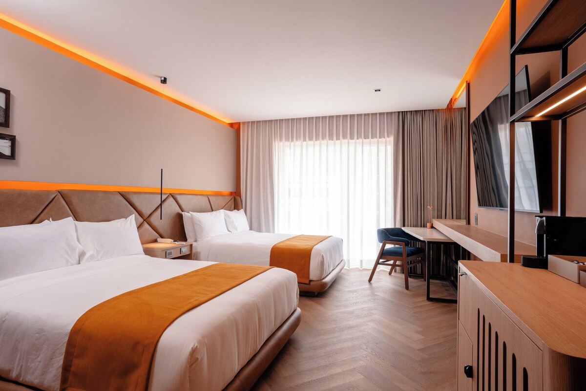 A hotel room with two beds, a desk, a chair, a TV, and a large window with curtains.