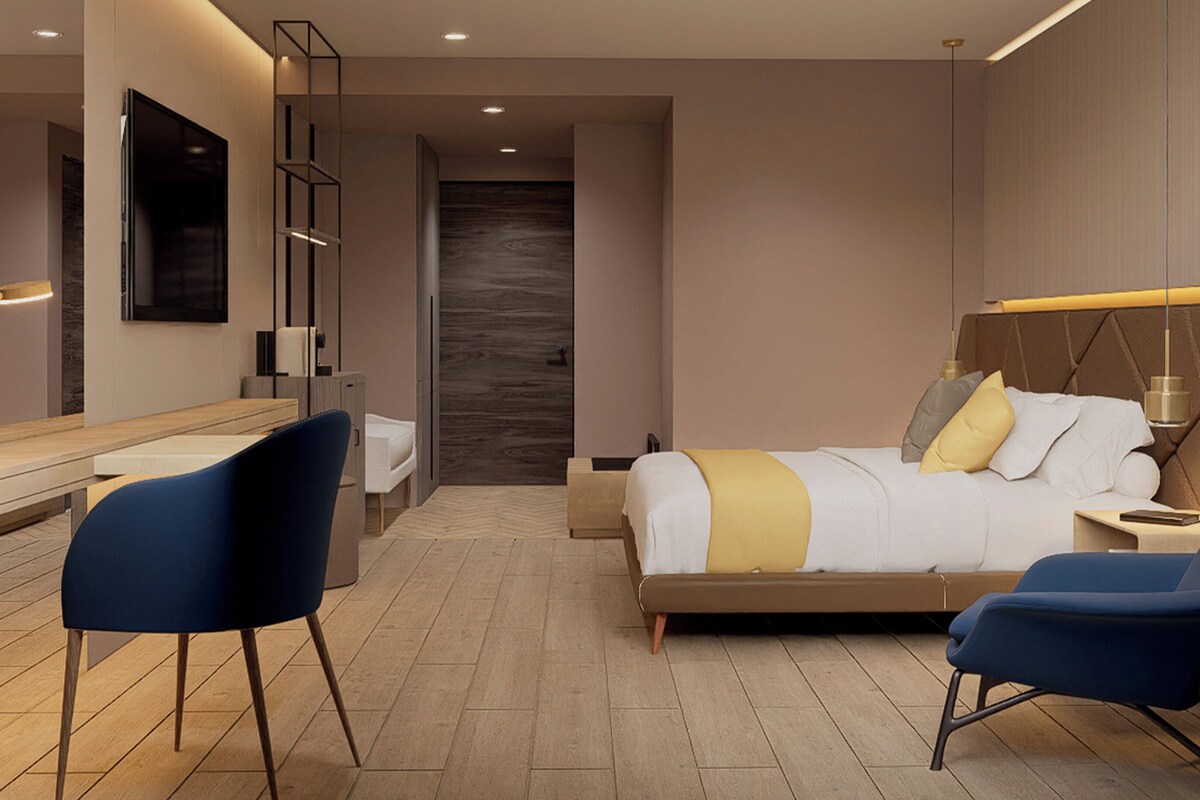 A modern hotel room with a bed, bedside table, chair, desk, and a large mirror.