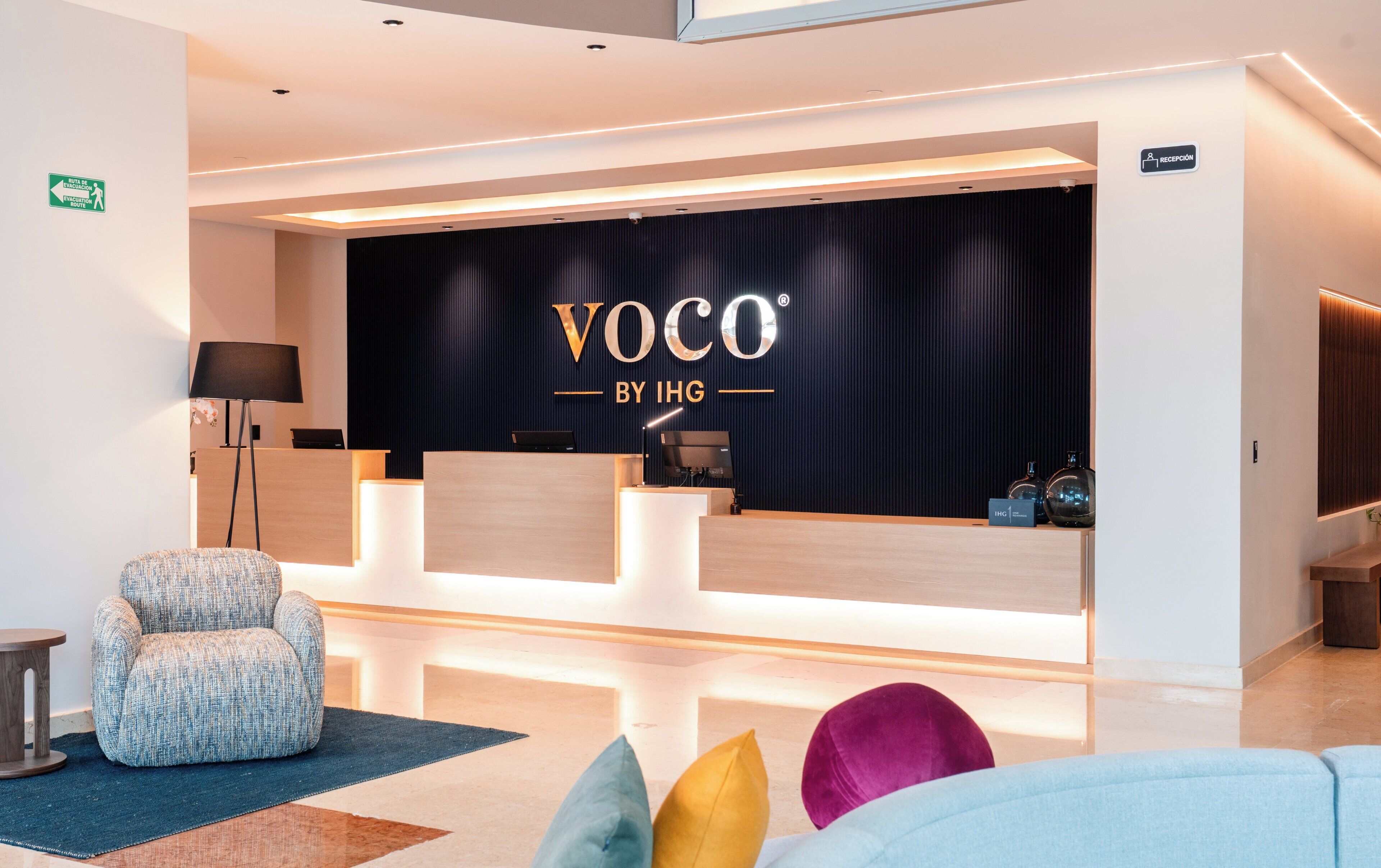 A reception area with a black backdrop displaying 'VOCO BY IHG' branding, a reception desk, a seating area with a patterned armchair, and a reception sign indicating 'RECEPTION'.