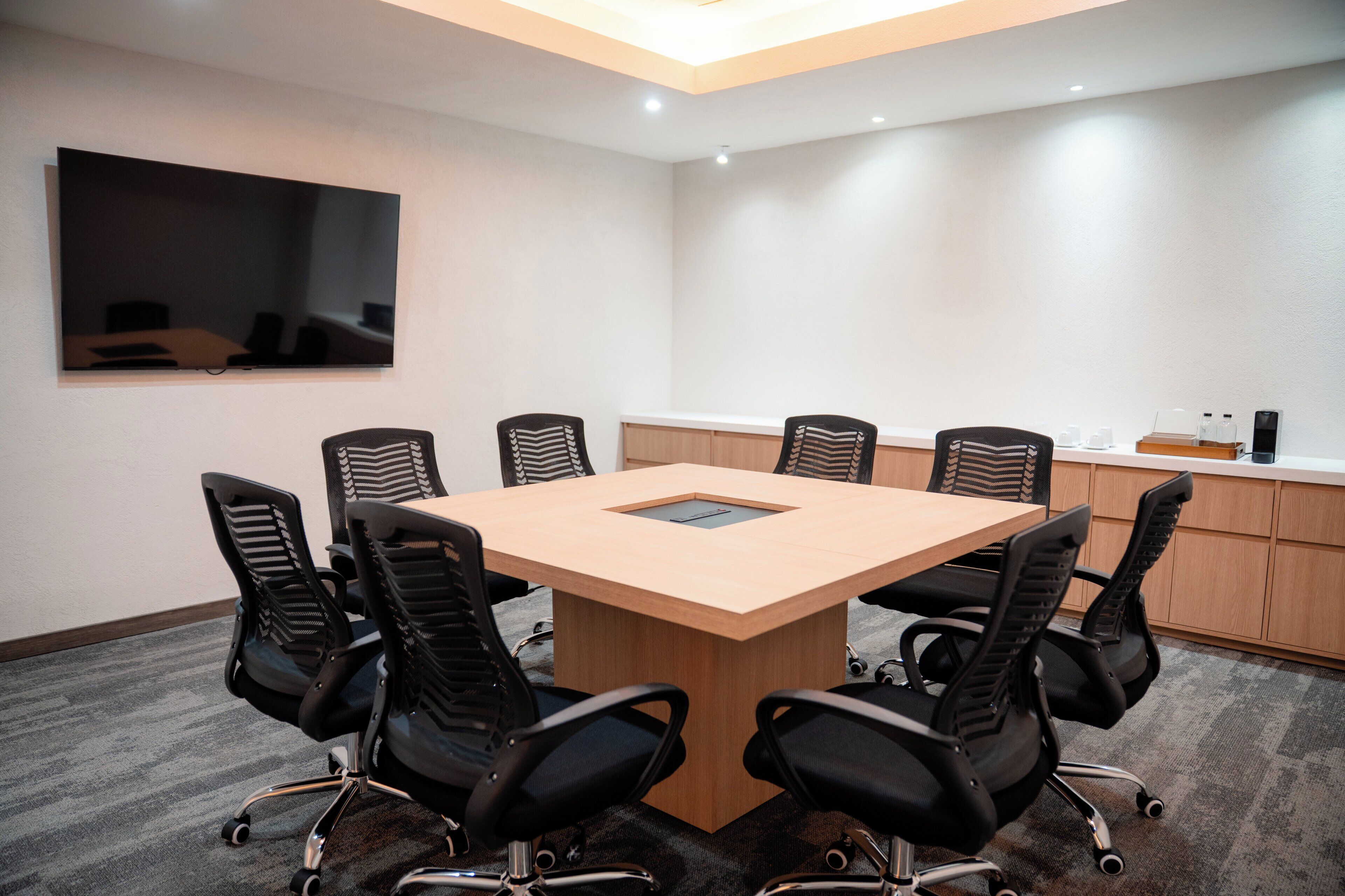 A conference room with a large table, six chairs, a flat-screen TV, and a kitchenette area.